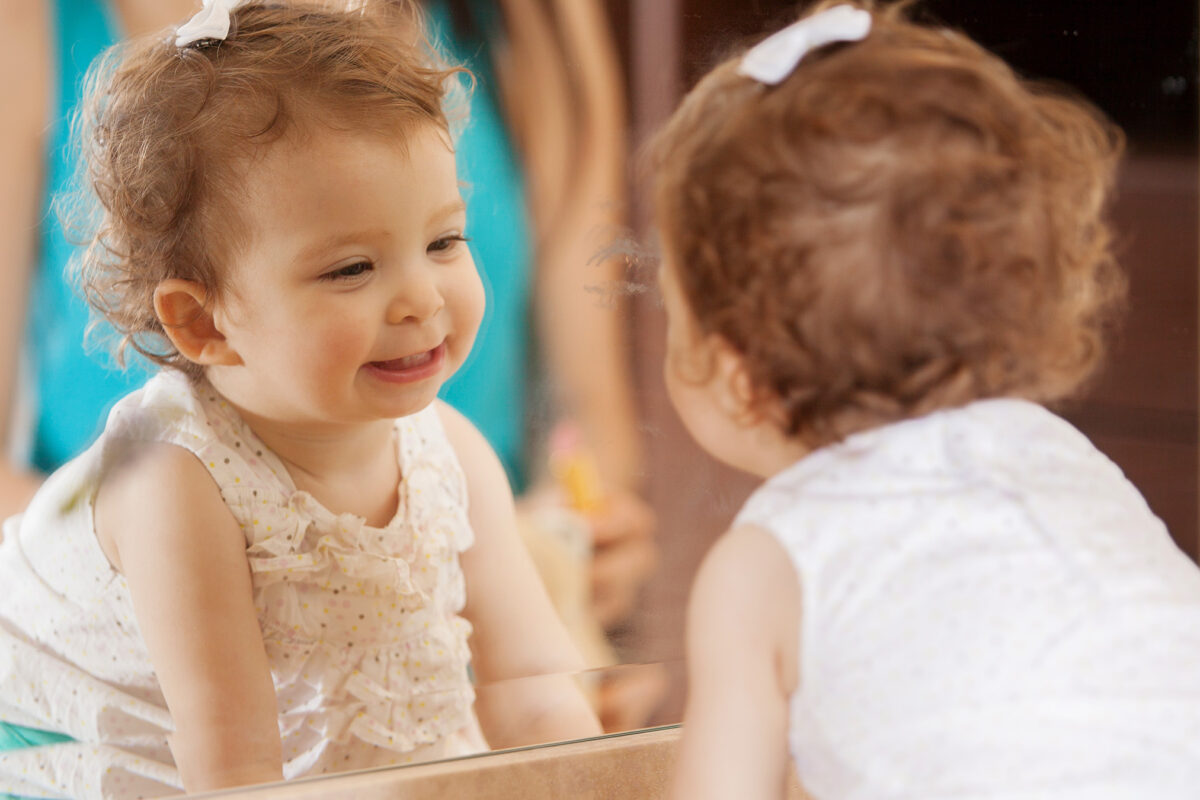 A toddler gazes at herself in the mirror and smiles.