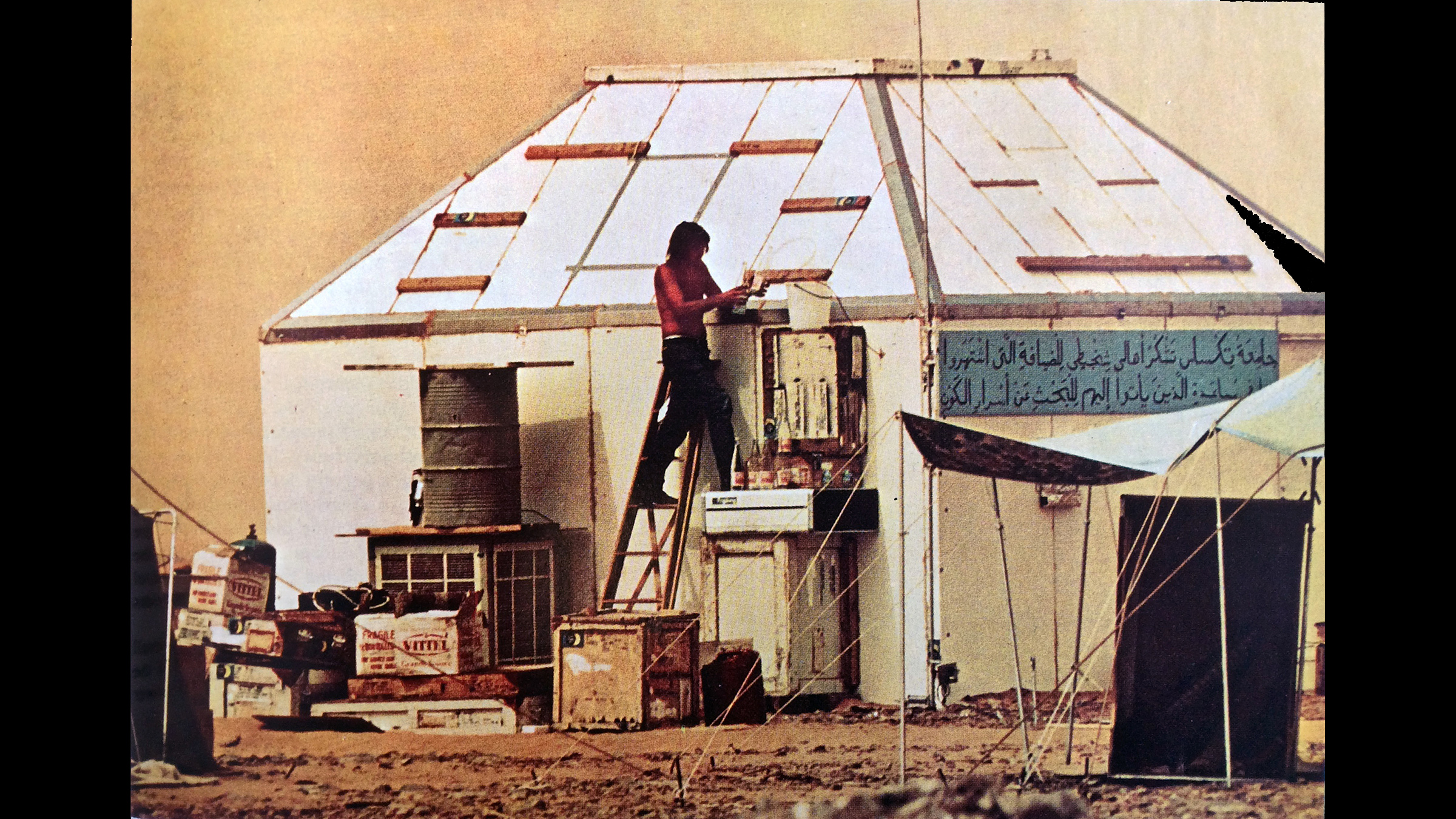 A man stands on a ladder outside a white hut in the desert