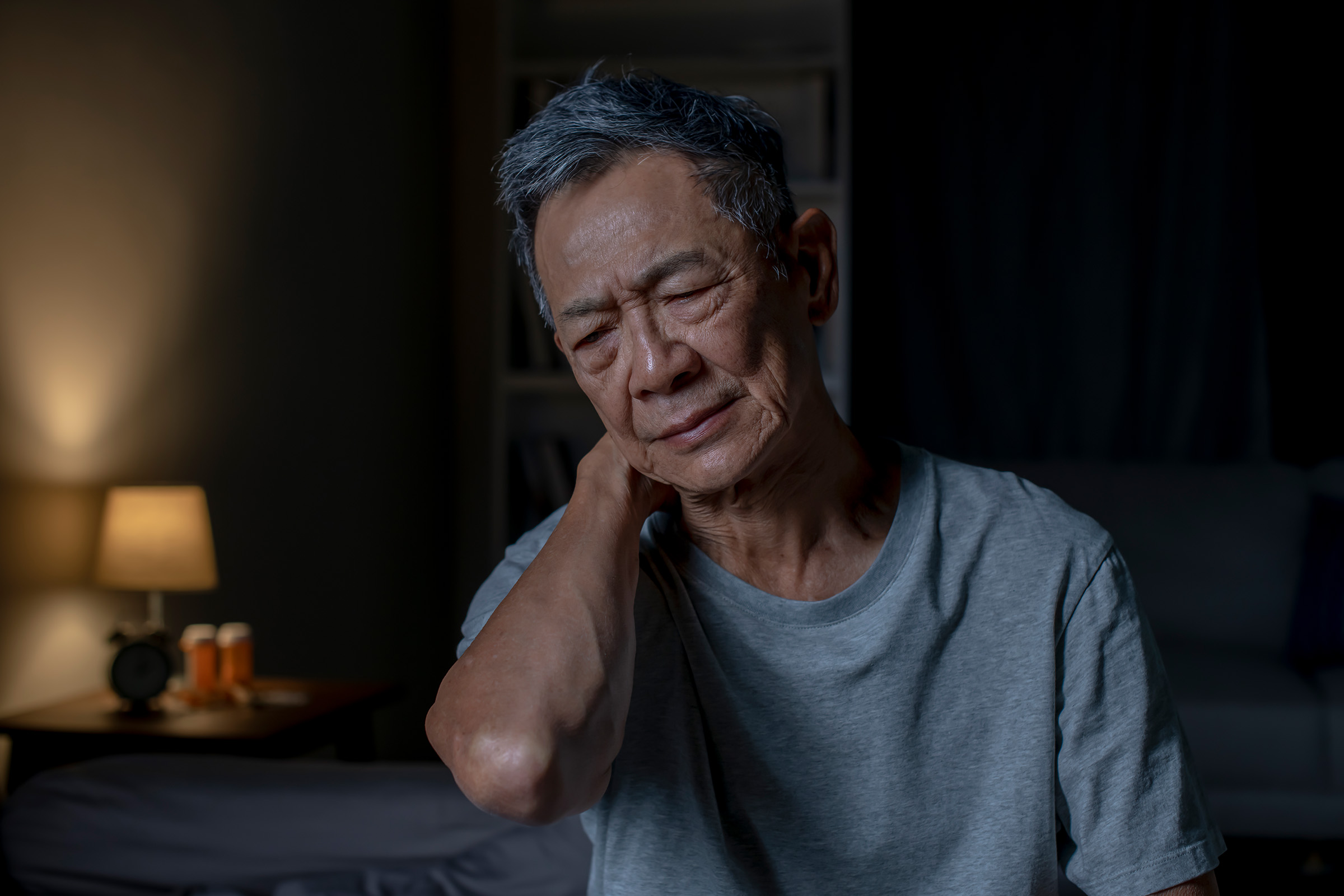 Older man sitting in dark room with hand on back of neck looking tired. 